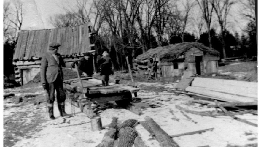 Man stands outside rustic cabins in a snowy rural setting.