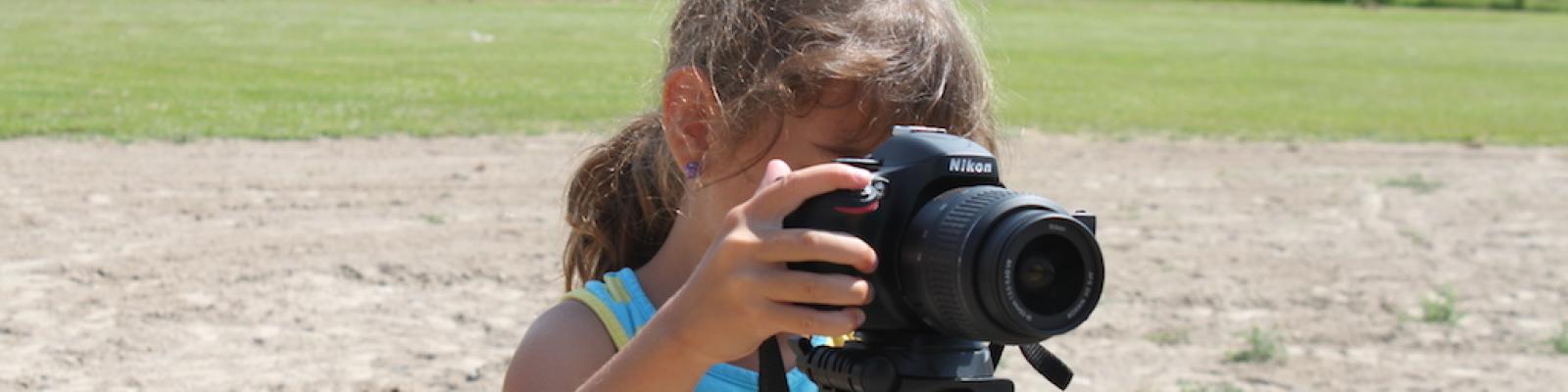Young girl in a dress using a camera on a tripod outdoors.