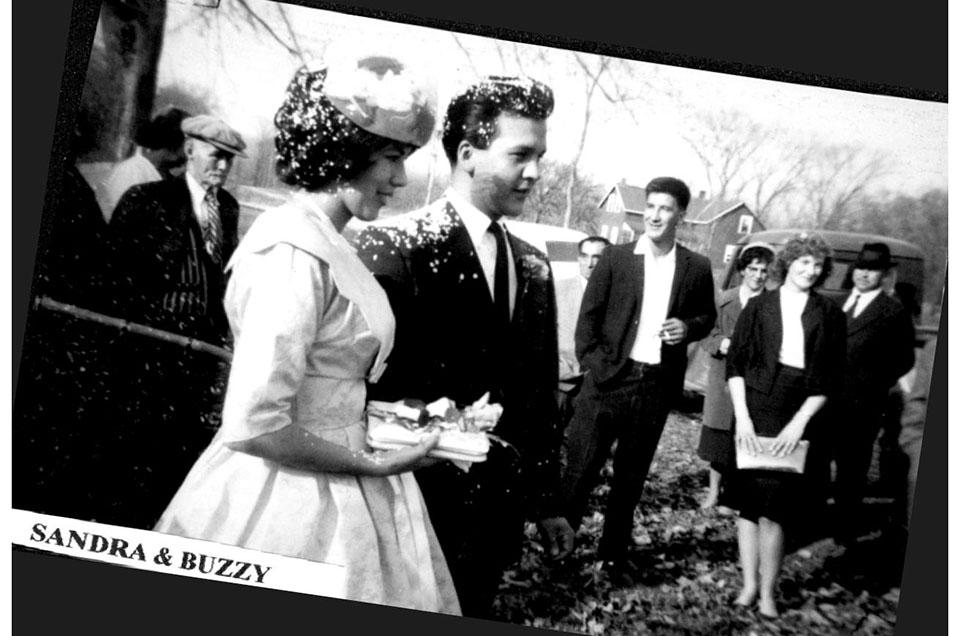 Black and white photo of a couple at an outdoor wedding, surrounded by smiling guests.