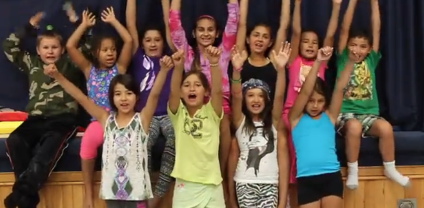 A group of kids sitting on a stage with their hands up in the air cheering. 