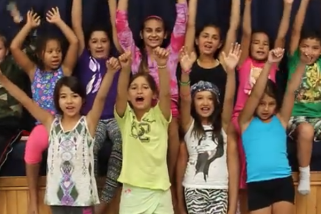 A group of kids sitting on a stage with their hands up in the air cheering. 