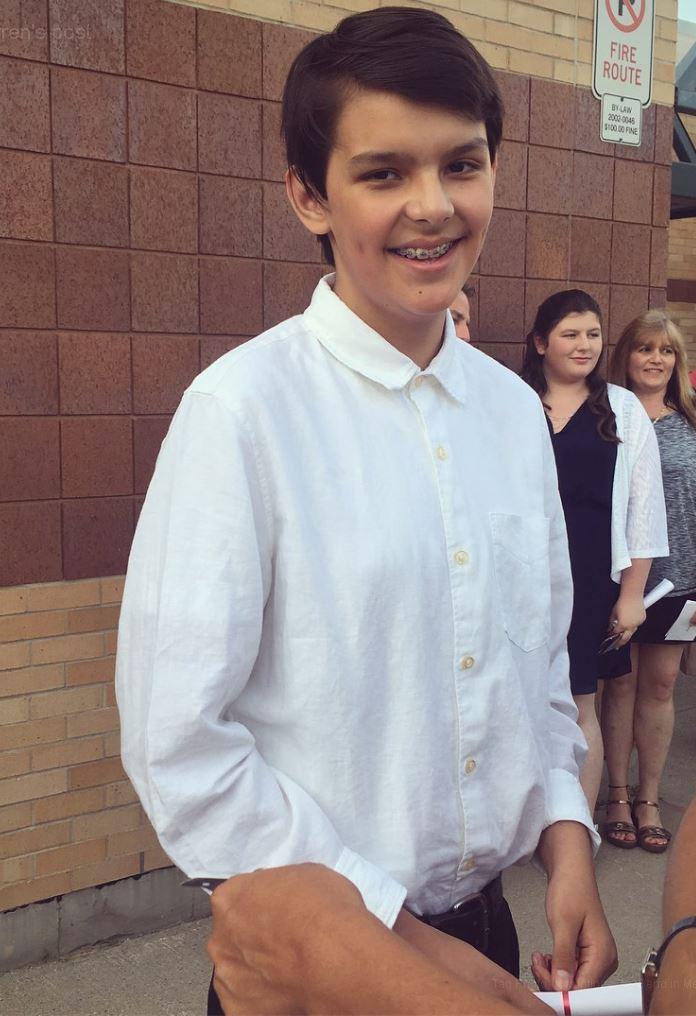 Young person in a white shirt smiling outdoors, with people in the background.