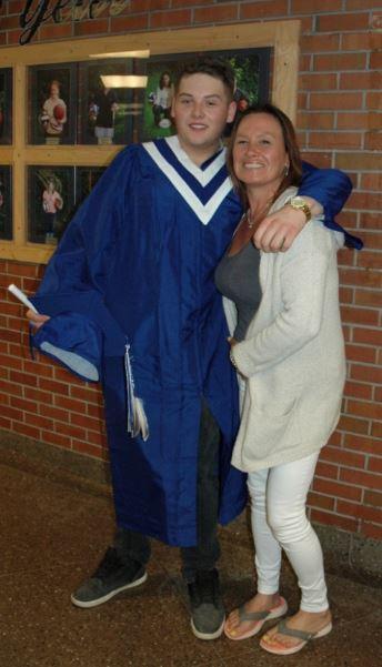 Graduate in blue gown hugs woman, both smiling by a brick wall indoors.