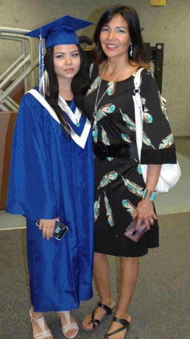 Graduate in blue gown and cap, standing with a woman in a patterned dress, both smiling.