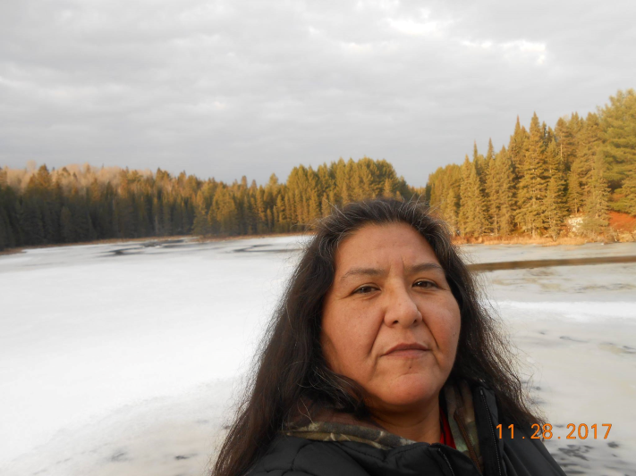 Woman by a frozen lake with pine trees in the background.