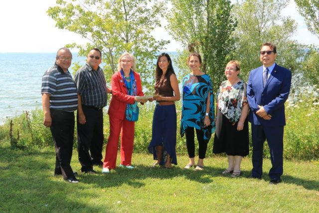 Seven people stand together on grass by a lake, under sunny skies.