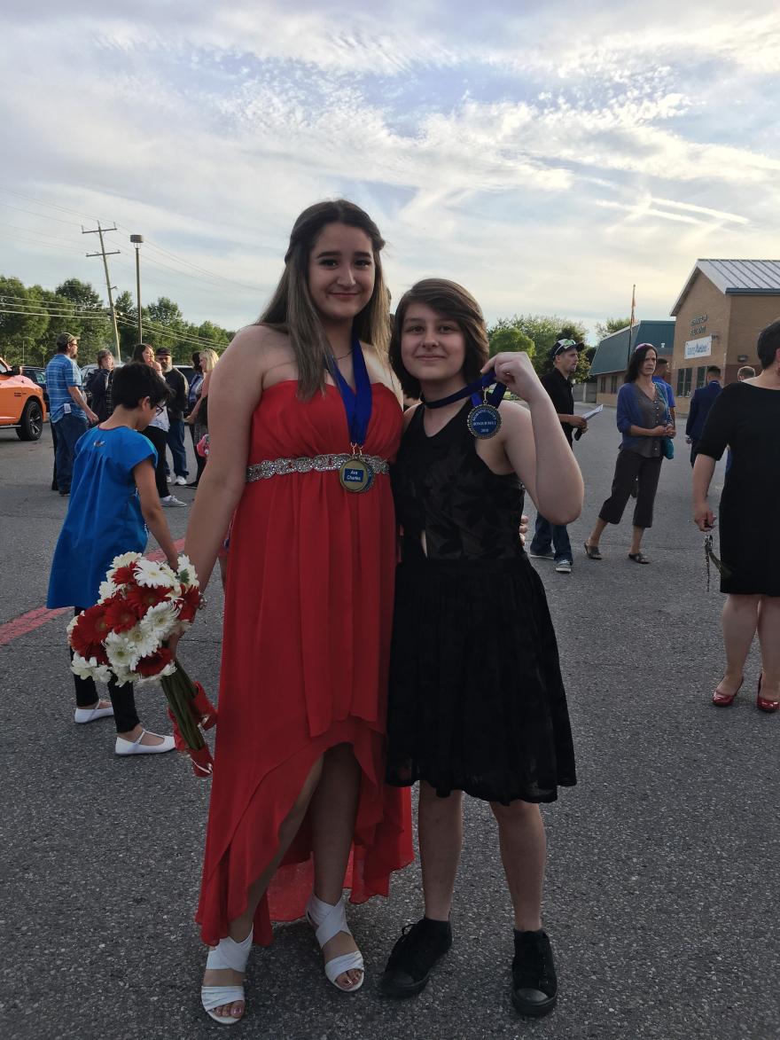 Two people smiling; one in a red dress holding flowers, the other in a black dress.