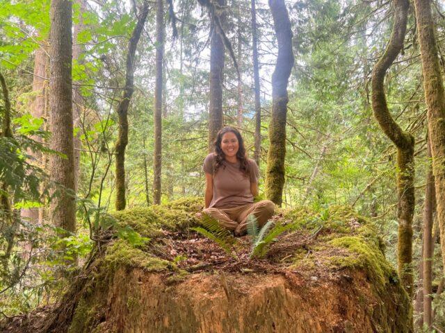 Woman sitting on a large mossy tree stump in a forest.