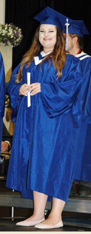 Graduate in blue cap and gown holding diploma, smiling on stage.