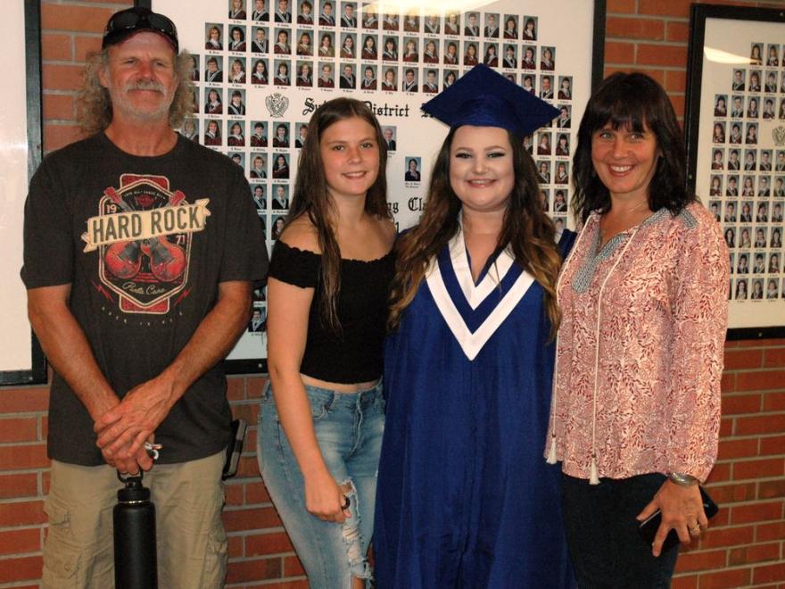 Family posing with graduate in blue gown and cap, smiling in front of photo wall.