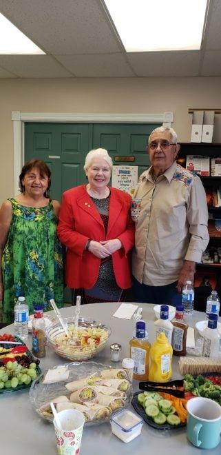 Three people standing behind a table with food in a room, smiling.