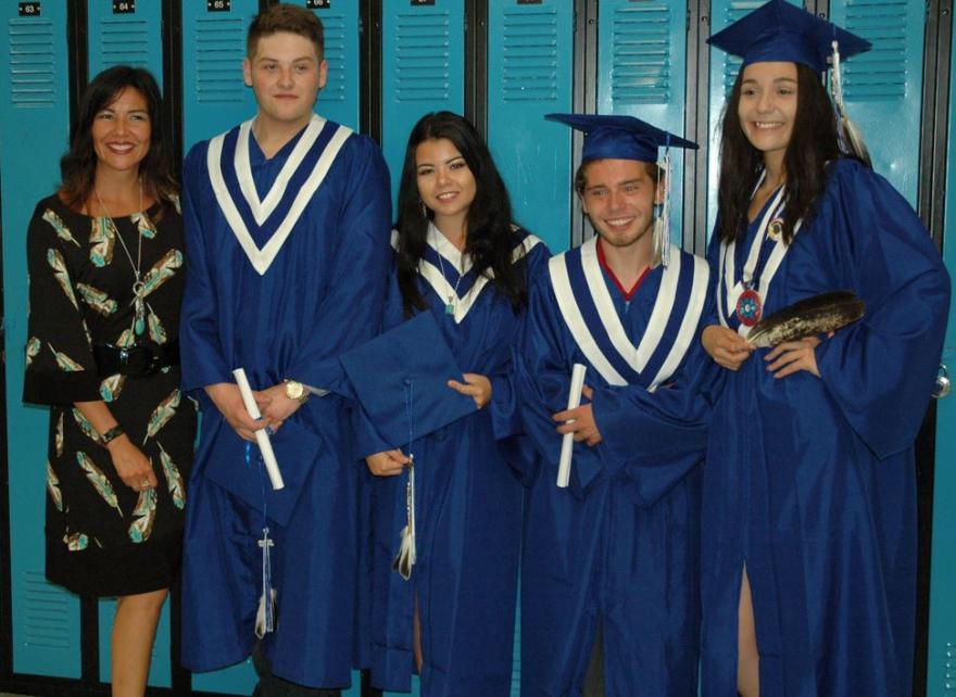 Graduates in blue gowns and caps pose with a teacher in front of lockers.